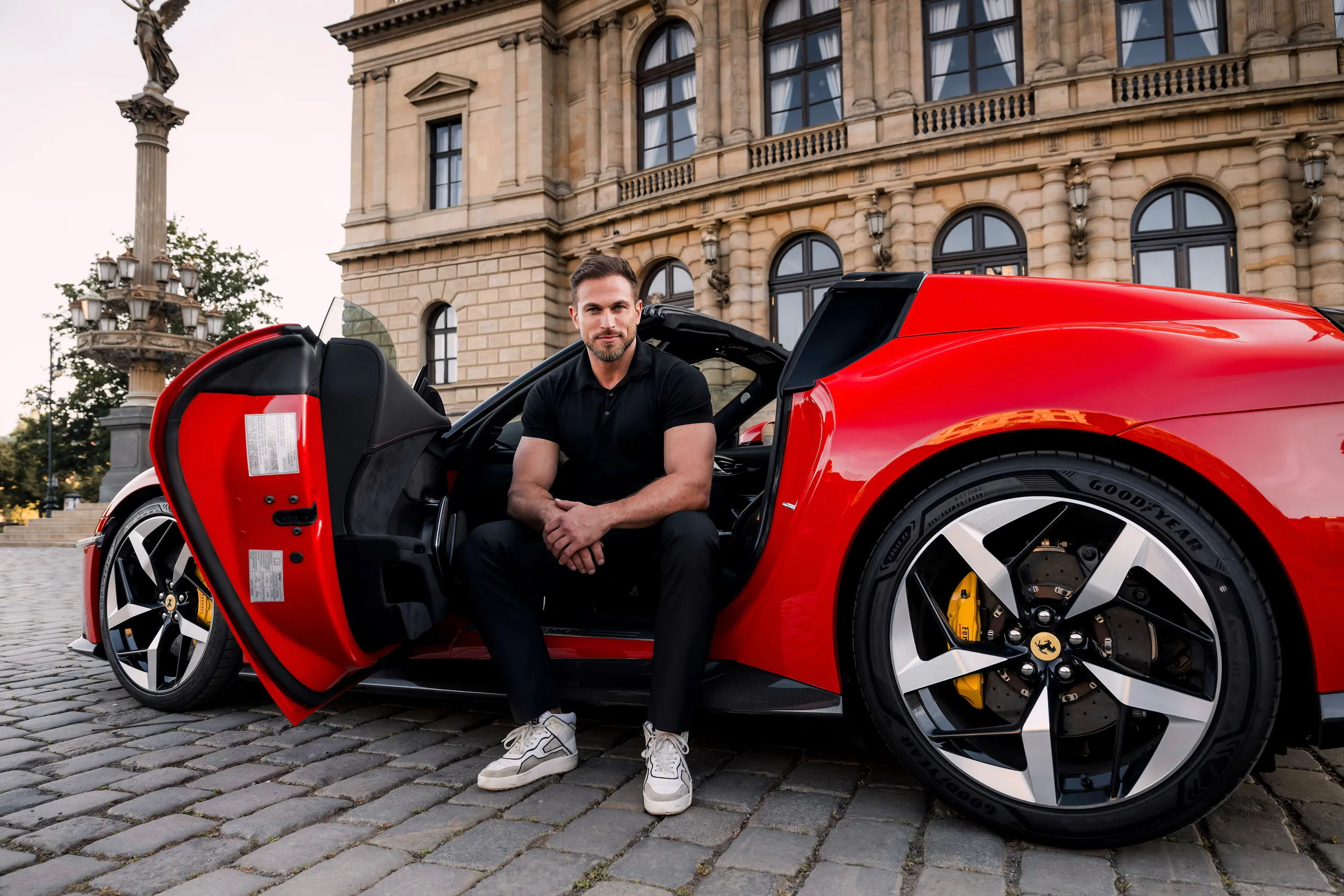 A man in black seated in the open door of a red Ferrari 12Cilindri Spider